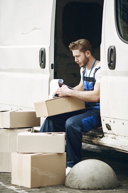 A man with a beard, wearing a blue and white striped shirt and dark blue work overalls, is sitting at the open rear of a white delivery van. He is holding a clipboard with papers and appears to be reviewing or organizing documents related to a home relocation or furniture transport. Surrounding him on the ground are several cardboard boxes, some open and some sealed, indicating packing or unpacking activities. Inside the van, visible behind him, are more boxes and packaging materials, including additional cardboard containers. The van is parked on a paved surface with a concrete bollard nearby, and daylight illuminates the scene, suggesting daytime. This image exemplifies a loading or planning phase in house removals, showcasing the careful handling and organization of packed items as part of a professional moving process, as offered by Man with Van Catford.