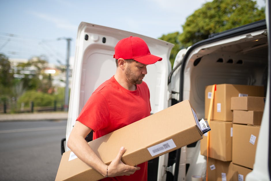 A man with a beard, wearing a blue and white striped shirt and dark blue work overalls, is sitting at the open rear of a white delivery van. He is holding a clipboard with papers and appears to be reviewing or organizing documents related to a home relocation or furniture transport. Surrounding him on the ground are several cardboard boxes, some open and some sealed, indicating packing or unpacking activities. Inside the van, visible behind him, are more boxes and packaging materials, including additional cardboard containers. The van is parked on a paved surface with a concrete bollard nearby, and daylight illuminates the scene, suggesting daytime. This image exemplifies a loading or planning phase in house removals, showcasing the careful handling and organization of packed items as part of a professional moving process, as offered by Man with Van Catford.