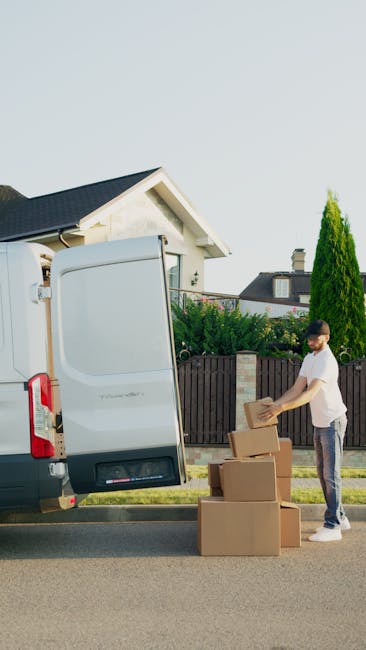 A man from Man with Van Catford is preparing for a home relocation on a residential street, standing beside a light grey moving van with its rear door open. He is wearing casual clothing, including a