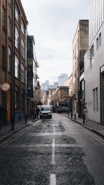 A man from Man with Van Catford is preparing for a home relocation on a residential street, standing beside a light grey moving van with its rear door open. He is wearing casual clothing, including a