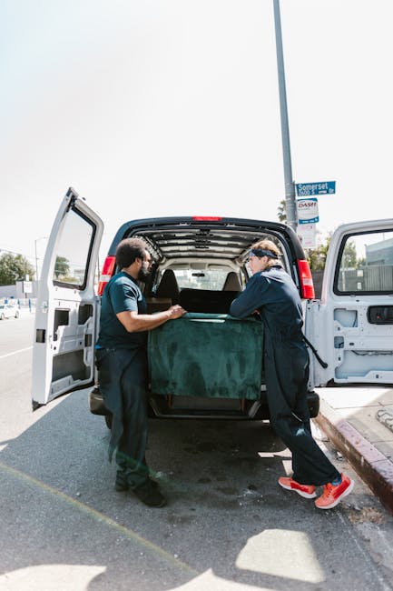 Two professional movers from Man with Van Catford are loading a large, dark green upholstered piece of furniture into the rear of a white panel van parked on a paved street. The furniture appears to b