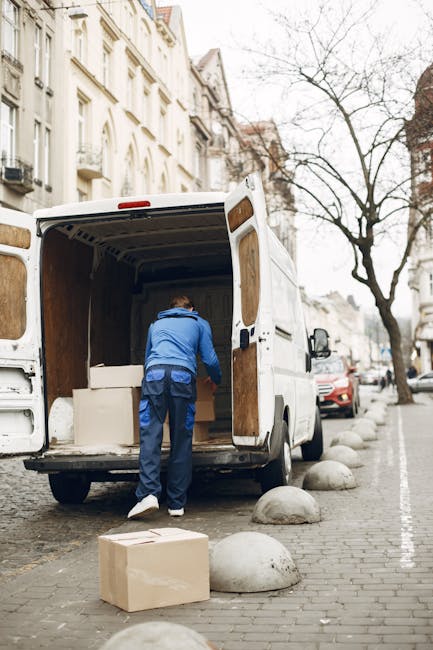 Two professional movers from Man with Van Catford are loading a large, dark green upholstered piece of furniture into the rear of a white panel van parked on a paved street. The furniture appears to b