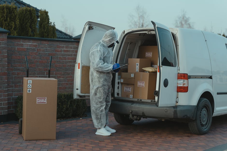 A person dressed in full protective PPE, including a white coverall suit, face shield, gloves, and mask, is standing next to a white commercial van with its rear doors open. The individual appears to 